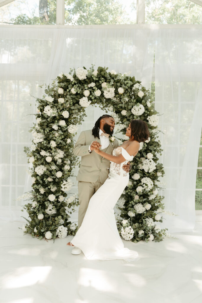 Black couple dressed in wedding attire doing a dip in front of a green and white arch inside a greenhouse. 