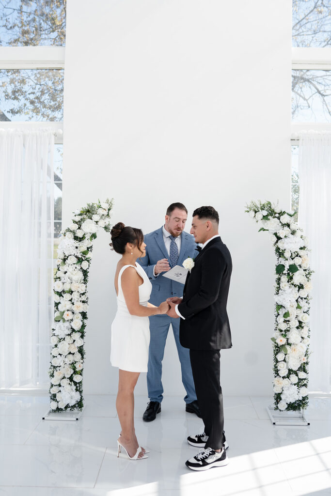 Bride and groom standing at The Makenzie House glass photo studio ceremony alter, in front of an officiant and a broken flower arch.